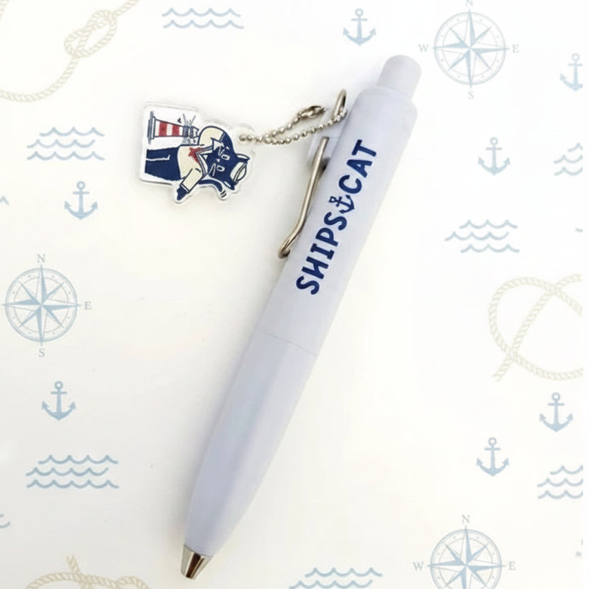 A white retractable pen with 'SHIPS & CAT' printed in navy blue, shown with a small lighthouse charm and metal keychain clip on a white background with faint nautical illustrations.