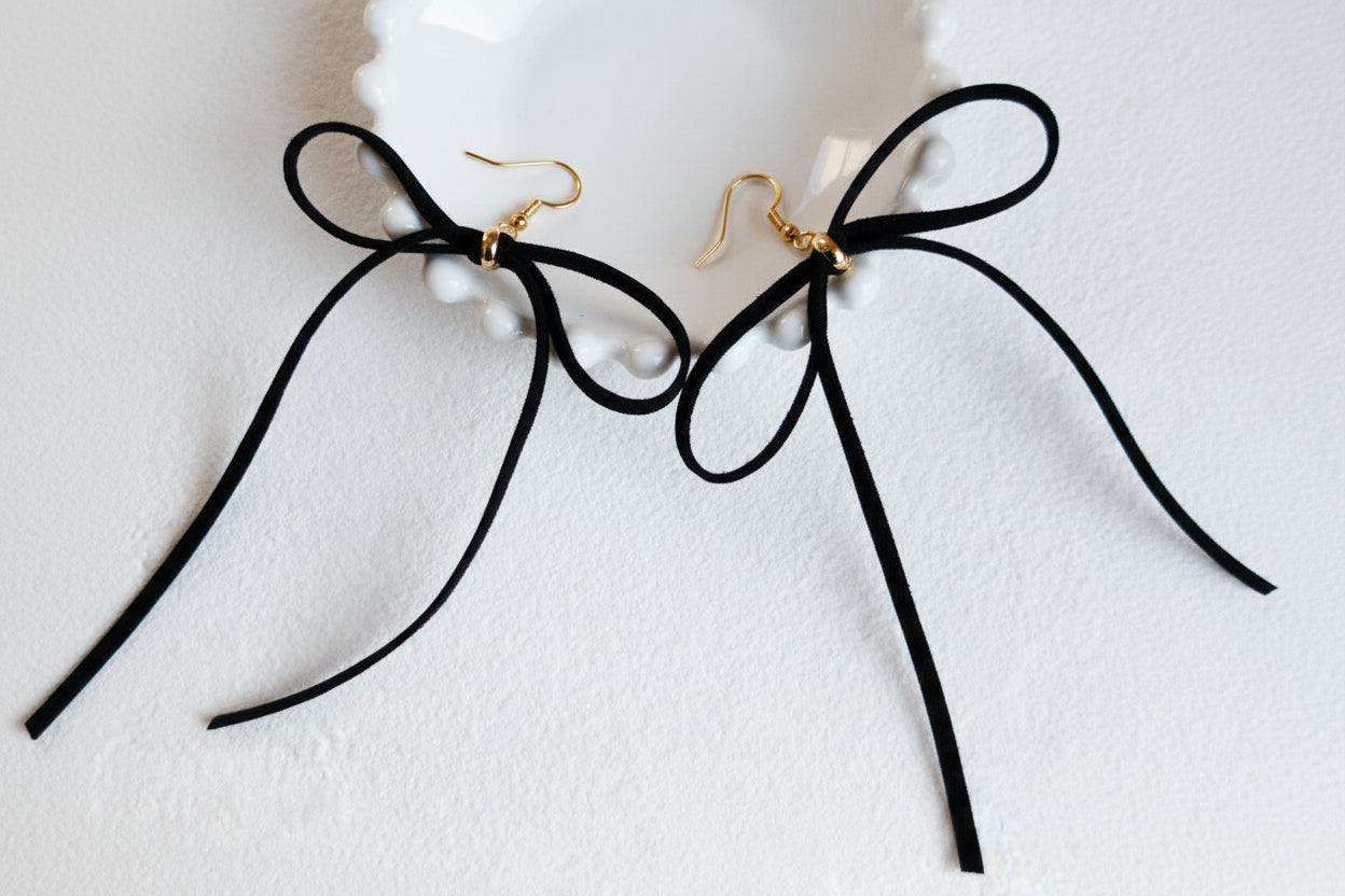 Black ribbon bow earrings with gold-tone clasp backs, displayed on a white scalloped dish against a white textured background.