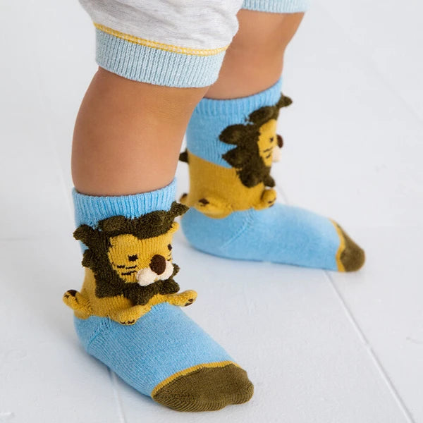A close-up of a child's feet wearing light blue socks with a 3D lion face design on the front, paired with white tights with a yellow stripe.