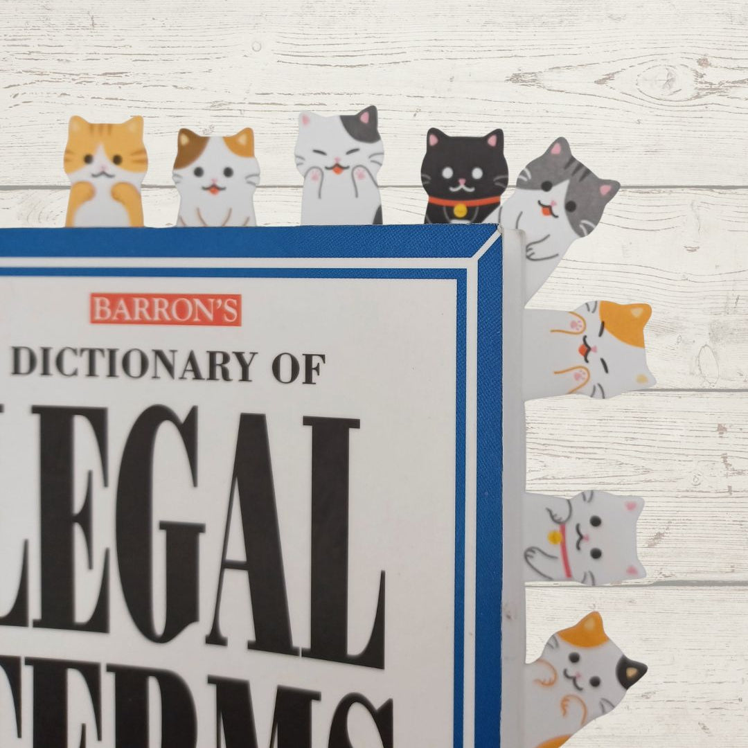 A close-up of a book titled 'Barron's Dictionary of Legal Terms' with colorful cartoon cat bookmarks attached along the right edge, set against a whitewashed wood background.