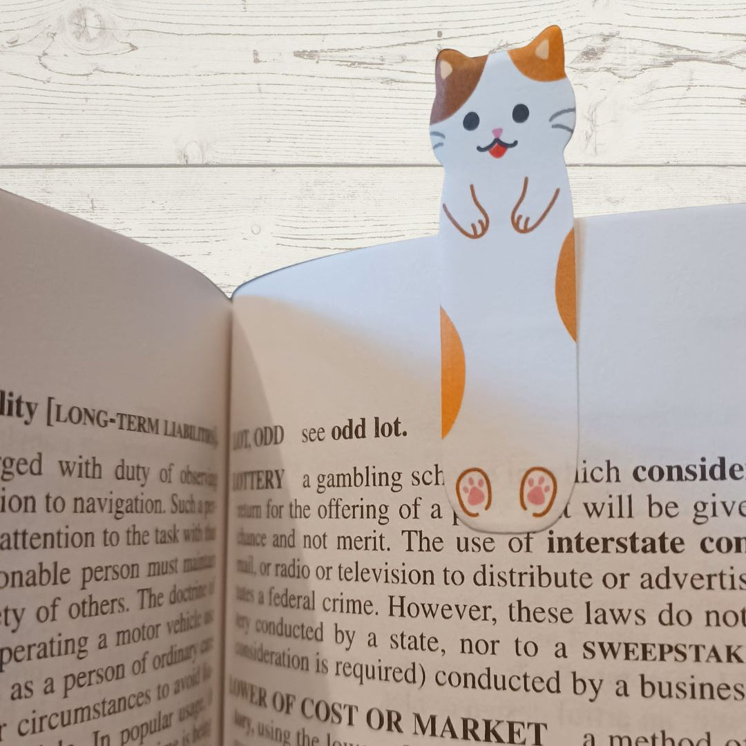 A decorative paper book bookmark shaped like a smiling orange-and-white cat peeking out from an open book.