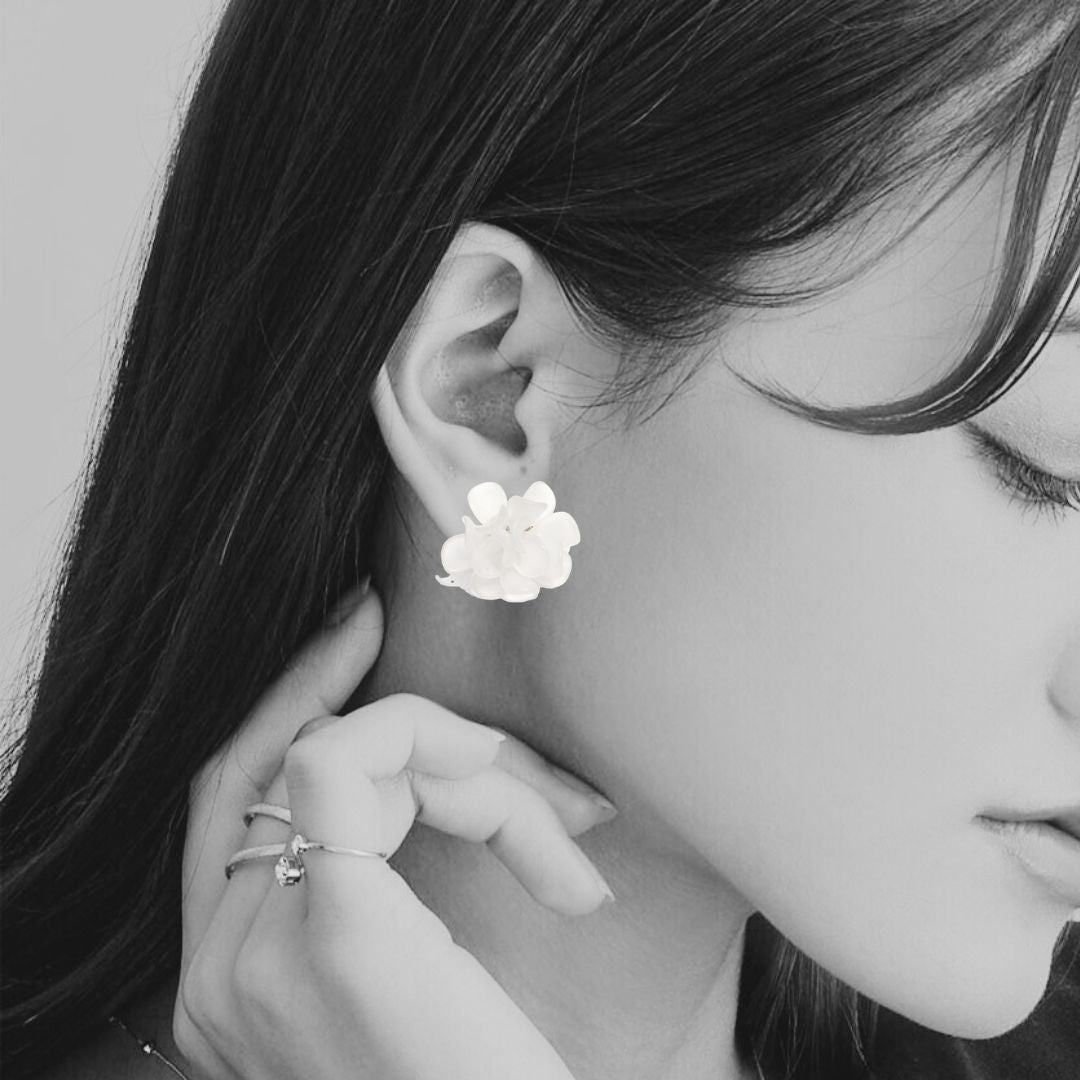 A close-up black and white photo of a woman wearing a small flower-shaped stud earring on her ear, with her hand near her neck and rings on her fingers.