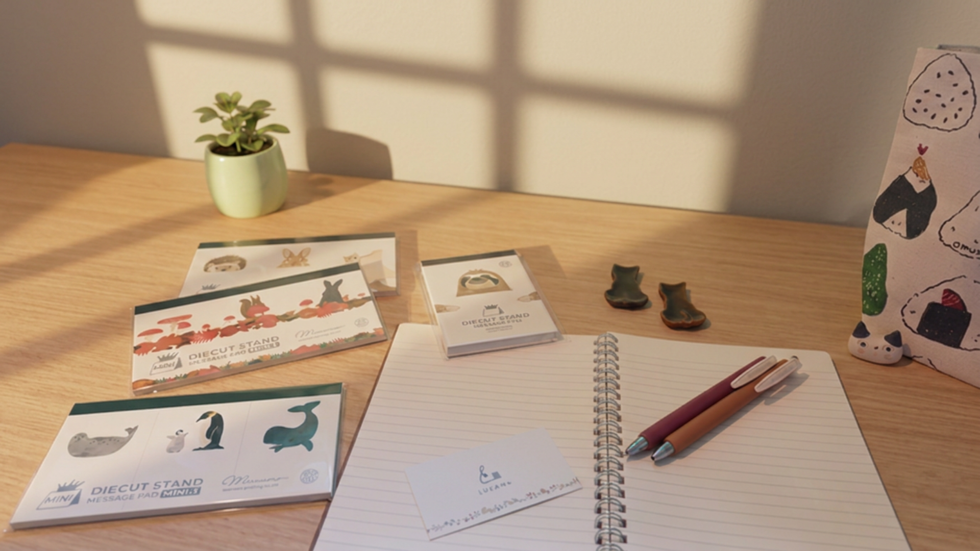A styled desk scene with printed stationery cards, a spiral notebook, two pens, small decorative items, and a patterned fabric bag on a wooden table. The scene is lit by warm window light with soft shadows cast across the surface.