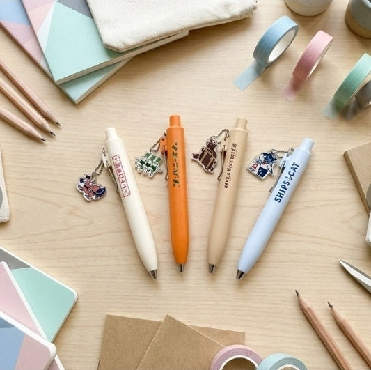 Four decorative ballpoint pens in cream, orange, beige, and light blue are arranged on a light wood desk with notebooks, washi tape, pencils, and small accessories around them. The pens have printed text and charm keychains attached.